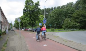 A bike path with two people on bikes beside a roadway that turns.